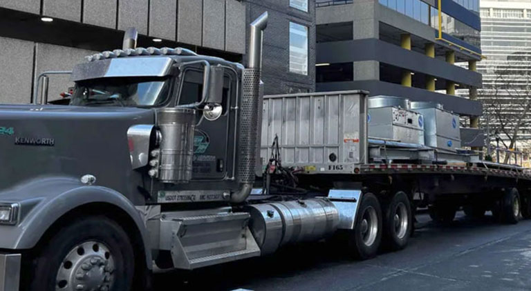 Heavy haul transport truck carrying commercial HVAC equipment past city buildings.