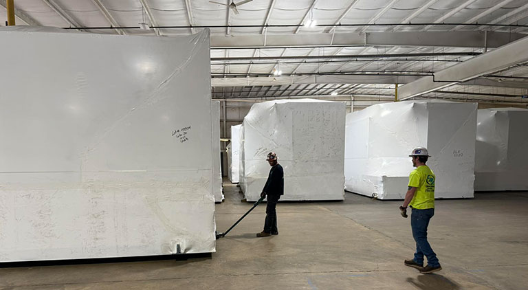 CMR Crane and Rigging workers positioning large shrink-wrapped machinery in a warehouse using heavy equipment skates.